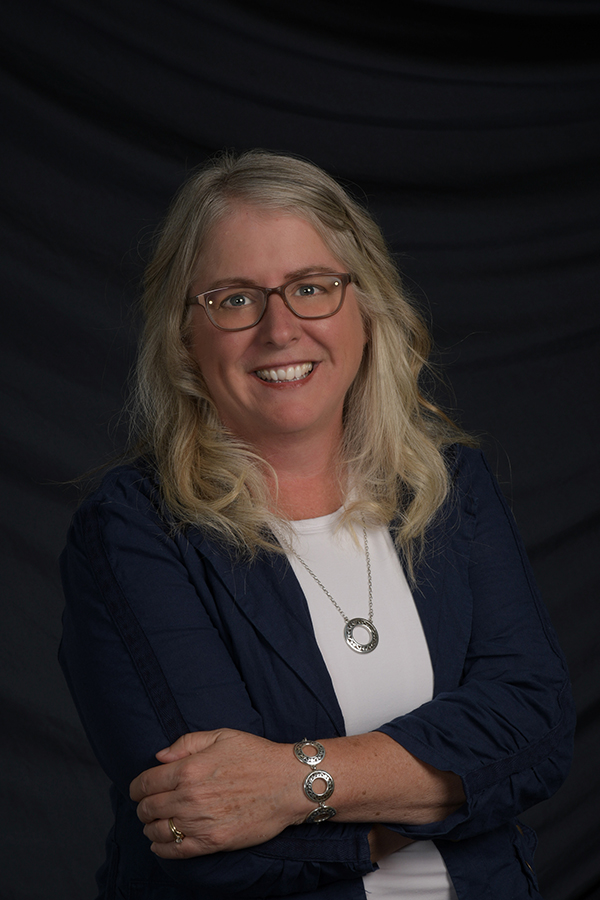 Smiling woman with long blonde hair and glasses, wearing a navy jacket over a white shirt, stands with arms crossed in front of a dark, draped background—exuding confidence and a commitment to lifelong learning.